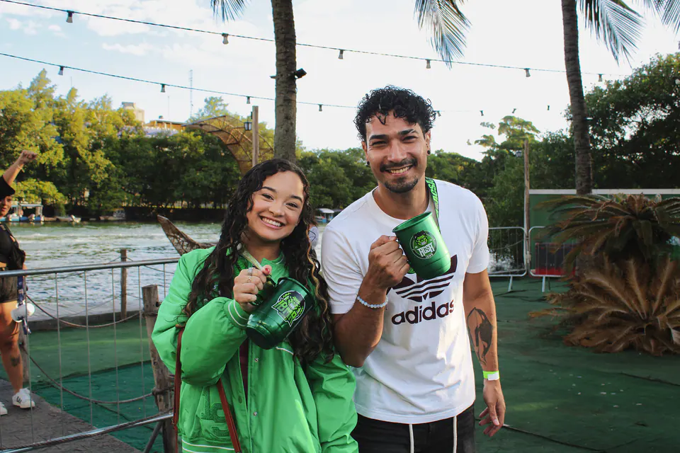 casal sorri à beira do rio segurando as canecas verdes da festa